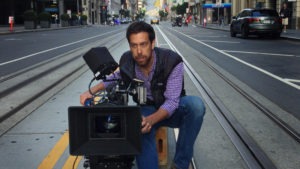 Dan Krauss crouches on a city street, operating a professional video camera. The street is empty, with tracks visible and a few vehicles and motorcycles in the background. Focused on his work, he wears a checkered shirt and vest, clearly on a roll capturing the perfect shot.
