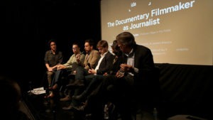 Six people sit on a stage in discussion under the title "The Documentary Filmmaker as Journalist" projected on a screen behind them. They appear to be part of a panel discussion at an event, exploring how journalism intersects with filmmaking in modern media.