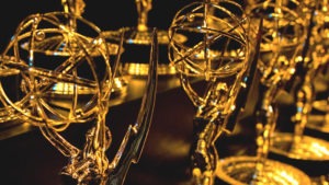 A close-up shot of multiple News and Documentary Emmy Awards statues in golden color. The trophies, featuring a winged woman holding an atom, stand on circular bases. Arranged in rows and shining brightly under the lighting, they glow against the dark background, reminiscent of a red carpet event for J-School Alumni.
