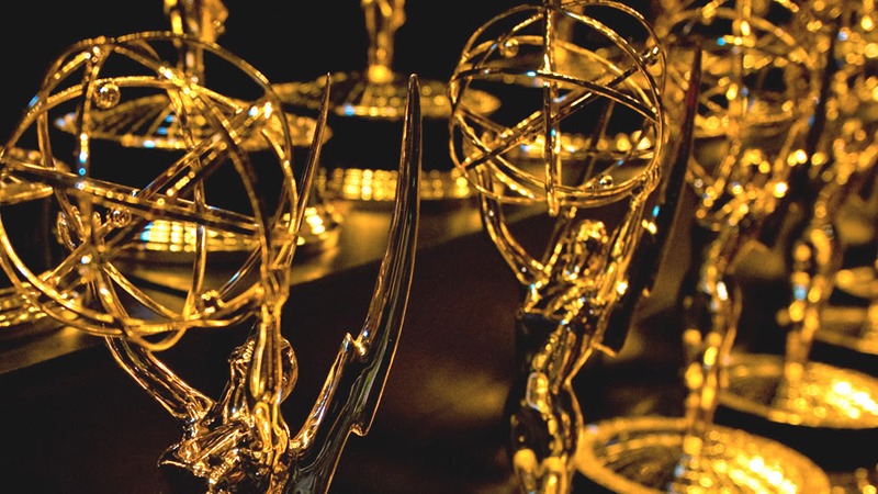 A close-up of several golden Emmy Awards trophies, featuring a winged woman holding an atom. The trophies are arranged in rows, reflecting a soft light that emphasizes their metallic shine. The background is dark, making the awards stand out prominently like stars in a news documentary.