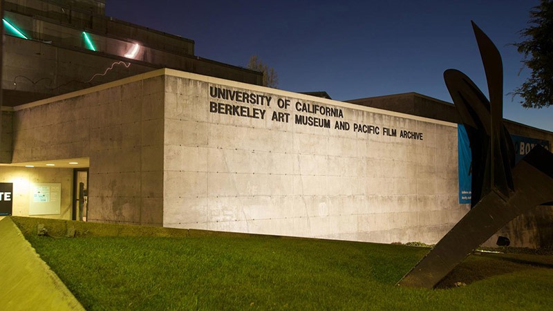 The image shows a nighttime view of the exterior of the University of California Berkeley Art Museum and Pacific Film Archive. The concrete building is illuminated, featuring a large sign with its name. In the foreground, part of a metal sculpture envisioned for future generations is visible on a grassy area.