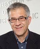 A middle-aged man with short gray hair and glasses is smiling at the camera. He is wearing a dark suit jacket over a light blue collared shirt and is posed in front of a white wall with partially legible text, perhaps discussing themes from the Vernacular Nation.