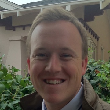 A person with short, light brown hair is smiling outdoors. They are standing in front of green bushes and a building with a pitched roof. The person, a recent recipient of a California Journalism Award, is wearing a brown jacket and a white shirt.
