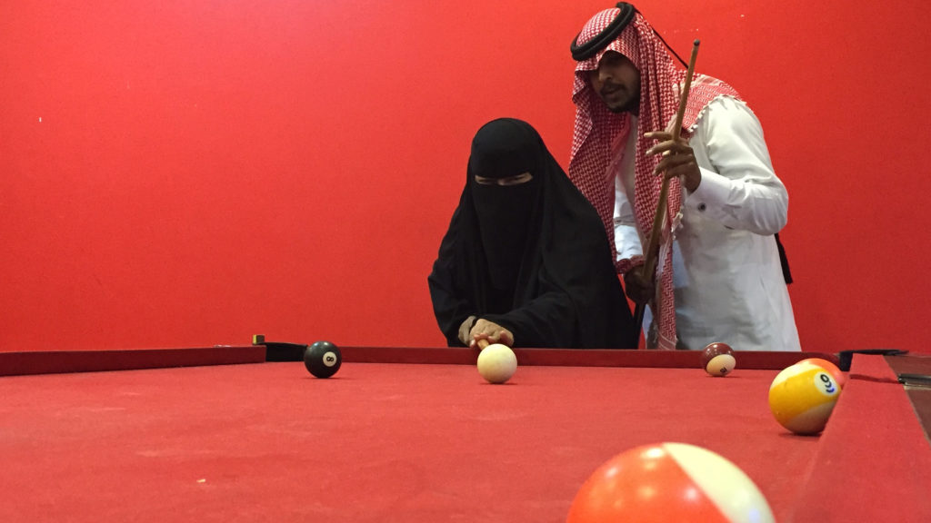 Two individuals are playing billiards. The person on the left is wearing a niqab and a black abaya, aiming a cue stick at the cue ball. The individual on the right, in a white thobe and red keffiyeh, is holding a cue stick, appearing to guide the player—a scene reflecting the changing face of Saudi women in sports.