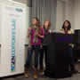 Three women, who appear to be students, are standing on a stage behind a podium giving a presentation. A banner to the left of the stage reads "NASA DEVELOP." They are dressed casually and seem to be in front of a lecture or classroom setting. It looks like part of a hackathon event.