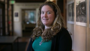 Harriet Rowan, a woman with long, wavy, light brown hair and a green top, smiles in a well-lit hallway. She is wearing a black cardigan and a necklace with a pendant. In the background are blurred wall photos and doors. Harriet recently gained national recognition for her work with Richmond Confidential.