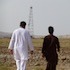 Two individuals, possibly including an IRP Fellow, are walking on a rocky terrain in Afghanistan towards a distant radio or cell tower. One person is dressed in white, and the other in brown. The sky is clear, and they appear to be in a remote, sparsely vegetated area.