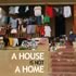 A house with a laundry line full of clothes hanging in front. The text "A House Is Not A Home" is written across the lower portion of the image, reminiscent of a J-School Student Book setup. A person sits on the stairs leading up to the entrance. The setting appears warm and lived-in.