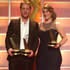 Two individuals in formal attire stand side by side holding awards. The recent grads on the left wears a dark suit and white shirt, while the person on the right dons a dark off-the-shoulder dress. They are on a stage with a bright, illuminated background.