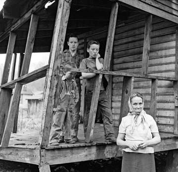 A black-and-white photo, evocative of a Berkeley Journalism piece, shows three children standing on the dilapidated porch of a wooden house while an elderly woman stands in the foreground. The children and woman have solemn expressions, and the porch is visibly worn and tilted.