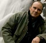 A person in an olive green jacket sits outdoors in front of a blurred nature background that appears to include a waterfall and rocks. The individual, possibly engrossed in deep thoughts on Berkeley Journalism, has a neutral expression and rests one arm on their knee.