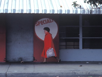 A person wearing a bright red coat walks on a sidewalk past a building with closed shutters and a "Special" sign in the background. The individual, perhaps rushing to an interview for Berkeley Journalism, carries a checkered bag and appears to be in motion. The setting looks urban and somewhat deserted.