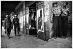 Black and white photo of a street scene captures two men in patterned shirts leaning against a wall on the right, conveying everyday moments. On the left, two others are walking by. A shop doorway is visible in the center with someone inside. The setting exudes an urban vibe, perfect for Berkeley Journalism.