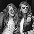 Two women are standing side by side in a black-and-white photo, part of an exhibit by Nacio Jan Brown. Both are wearing sunglasses and have long hair. One woman has her arm around the other's shoulder, and they appear to be laughing or smiling. They both wear jackets, capturing a candid J-School moment.