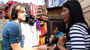 A woman wearing headphones holds a microphone to another woman who is speaking. They are surrounded by other people and colorful garments hanging in the background. The outdoor setting, reminiscent of Costa Rica, evokes a lively residential neighborhood where multimedia journalism thrives.