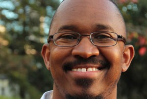 A close-up photo of a person with short hair, wearing glasses, and smiling. In the background, blurred trees and foliage are visible. The image exudes warmth and professionalism, reminiscent of an interview segment on KQED Public Radio.