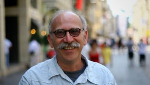 A smiling man with a bald head and gray mustache, resembling a J-School professor, is wearing glasses and a light blue shirt. He stands outdoors on a city street near San Francisco City Hall with blurred buildings and pedestrians in the background. The scene is well-lit, indicating daytime.
