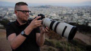 A man wearing glasses and a black shirt is outdoors, holding a DSLR camera with a large telephoto lens. He appears to be adjusting the camera settings, perhaps honing his skills like many J-School students who aspire to win top scholarships. The background shows an expansive cityscape under a hazy sky.