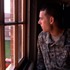 A person wearing a military uniform sits near a window, looking outside. The blurred background shows brick buildings and a green lawn. Warm light filters in through the window, casting a soft glow, reminiscent of scenes from Dan Krauss's films showcased at the Tribeca Film Festival.