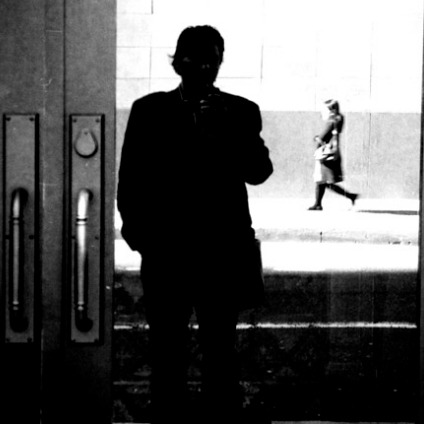 A black and white image shows the silhouette of a person capturing a photo in a reflective surface, resembling an Instagram-worthy moment. The reflection captures vertical handles and a woman walking on the sidewalk in the background. The scene has high contrast with distinct light and shadow areas.