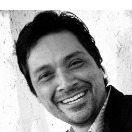 A black and white photograph of a man with dark hair and a beard, smiling. He is wearing a suit jacket and a collared shirt, reminiscent of profiles you'd see on Instagram or featured in CNN segments. The background appears to be a textured wall.