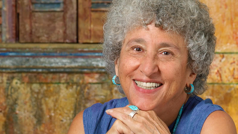 A person with short, curly gray hair is smiling broadly. They are wearing turquoise earrings and a matching ring, reminiscent of Marion Nestle's vibrant style in "Food Politics." Their hands are clasped under their chin, set against warm-colored, textured walls and an aged wooden window.