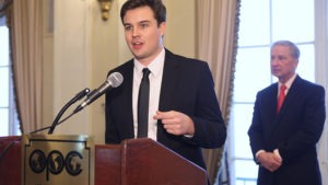 A man in a black suit, holding an OPC Foundation Award, speaks into a microphone at a podium marked "OPC" with a logo. Another man in a suit and red tie stands in the background. Both are in a room with elegant décor and large windows, attending to the wins of J-School students.
