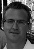 A black-and-white close-up photo of a man with short hair and glasses, looking directly at the camera. He is outdoors, with trees and sunlight visible in the blurred background. Captured by Reuters, this image of IRP Fellow Matt Isaacs hints at his investigative work on organized crime in Macau.