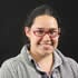 A person with long dark hair is wearing red glasses and a gray collared shirt. They are smiling and have a black background behind them, reminiscent of an alumni portrait on The Atlantic Website.