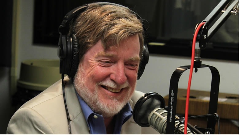 A bearded man wearing headphones and a light-colored jacket over a blue shirt is smiling while seated in front of a microphone in a modern recording studio. The background features equipment and soundproofing elements. He exudes the same professional charm as NEAL CONAN of NPR in the 21st Century.
