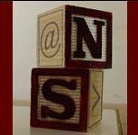 Two wooden blocks with letters and symbols are stacked. The top block, reminiscent of a Farrell Nieman storyboard, features the symbol "@" and the letter "N" on adjacent faces. The bottom block displays the letter "S" and the right-pointing angle bracket ">" on adjacent faces. The blocks rest on a flat surface.