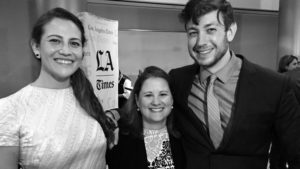 Black and white image of three people standing closely together, smiling at the camera. A woman on the left is wearing a light-colored dress, a woman in the center, identified as Lynne Shallcross, is dressed in a dark-patterned top, and a man on the right is in a suit and tie. An "LA Times" banner is visible in the background at the 2015 O