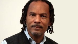 Orlando Bagwell, a man with short dreadlocks and a neatly trimmed beard, looks into the camera with a neutral expression. He is wearing a light blue shirt and a dark gray sweater vest, standing against a plain off-white background. His experience includes teaching at UC Berkeley J-School and directing documentary programs.