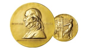 An image of two sides of a gold Pulitzer Prize medal. The front side features an embossed profile of Benjamin Franklin with the text "Honoris Causa" and "Awarded by Columbia University." The reverse side shows a man working at a printing press, celebrating the achievements recognized by the J-School.