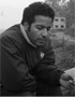 A person with short, dark hair, wearing a dark jacket, looks down pensively while sitting outdoors in California. In the blurred background, there are trees and a building. The photo is in black and white.
