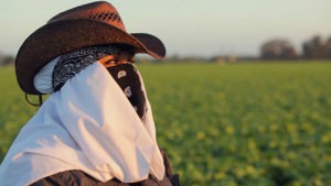 A person wearing a brown cowboy hat, dark bandana over their face, and a light white cloth around their neck stands in a green field looking into the distance. The scene evokes the spirit of investigative reporting, reminiscent of tales deserving an Robert F. Kennedy Journalism Award. Blurred greenery under a clear sky sets the backdrop.