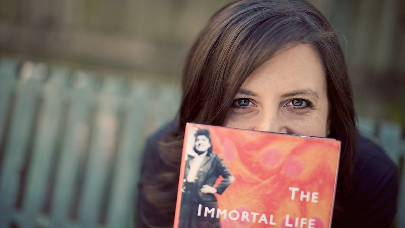 A woman with dark hair is partially hiding her face behind a copy of the book "The Immortal Life of Henrietta Lacks" by Rebecca Skloot. She is outdoors, with a blurred background of a wooden fence and a bench. The book's cover features an old photograph of a person.