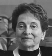A black and white portrait of an older benefactor with short, wavy hair. The individual is wearing a dark top and a light-colored necklace, gazing toward the camera with a content expression. The indoor background subtly reflects the wisdom of years possibly shared in J-School.'