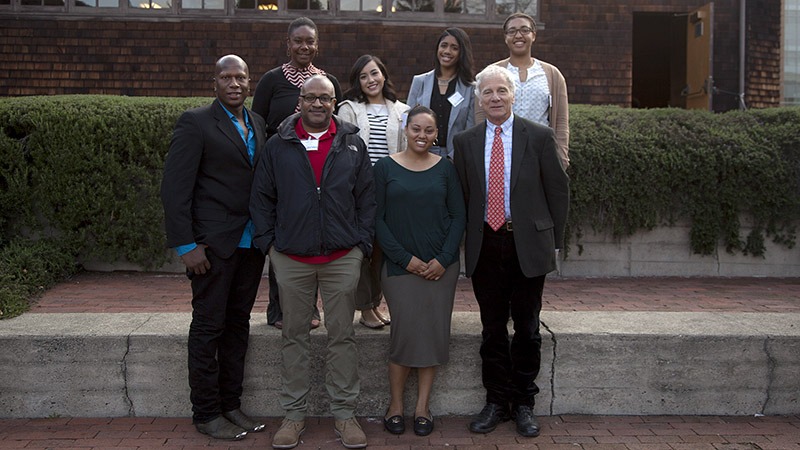 A group of eight diverse individuals, including Johnathan Rodgers, stands together outside, smiling for the camera. Dressed in a mix of business and casual attire, these fellows from J-School pose in front of a building with a window and greenery along a stone-edged walkway.