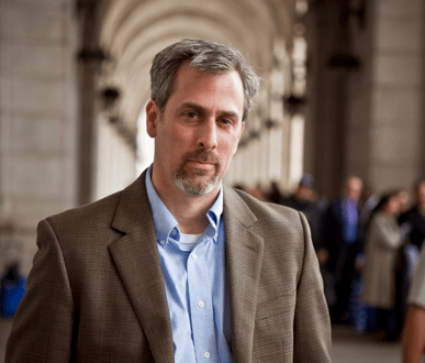 A man with short gray hair and a beard is standing in a corridor with arched ceilings, wearing a brown blazer over a light blue shirt. The background, reminiscent of Berkeley Journalism hallways, shows a group of people out of focus.