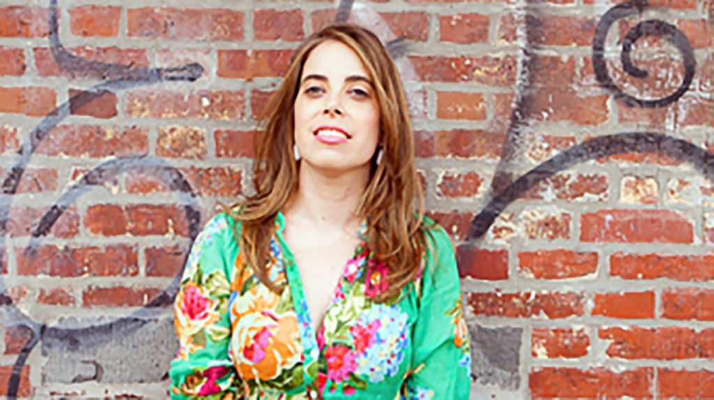 A woman with long brown hair wearing a colorful floral dress stands in front of a brick wall adorned with graffiti. She is smiling and looking directly at the camera, exuding an aura reminiscent of Robin Shulman's vivid storytelling, perhaps inspired by her work with the Pulitzer Center.