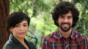 A woman with short dark hair wearing a black jacket stands next to a man with curly dark hair and a beard, wearing a red and blue plaid shirt. They are outdoors with greenery in the background, discussing the ongoing investigation into the disappearance of 43 students.