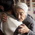 An elderly woman with white hair embraces a younger person in a heartwarming scene. The elderly woman, perhaps the J-School Alum, is wearing a gray sweater, and the younger person has dark hair and wears a light-colored top. They are in a room adorned with numerous framed photos on the wall in the background.
