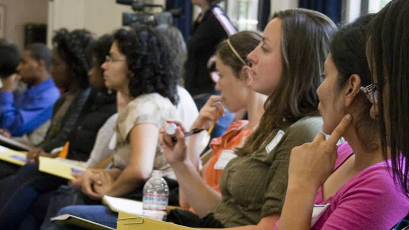 A diverse group of people sit in a row of chairs, attentively listening and taking notes during a lecture by Suzanne Franks on Women in Journalism. Some are holding yellow notepads and pens, while others have bottled water. A camera is visible in the background.