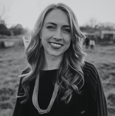 Maggie Beidelman, with long, wavy hair, smiles at the camera. She's wearing a dark top and a long necklace with several strands. The background is an outdoor setting with grass, trees, and blurred objects, creating a serene atmosphere. The image is in black and white.