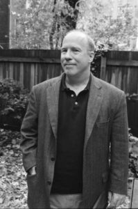 Mark Danner stands outdoors in a garden with a wooden fence behind him. He is wearing a dark blazer over a black collared shirt and has his hands in his pockets. The ground is covered in fallen leaves, and there are trees and shrubs around.