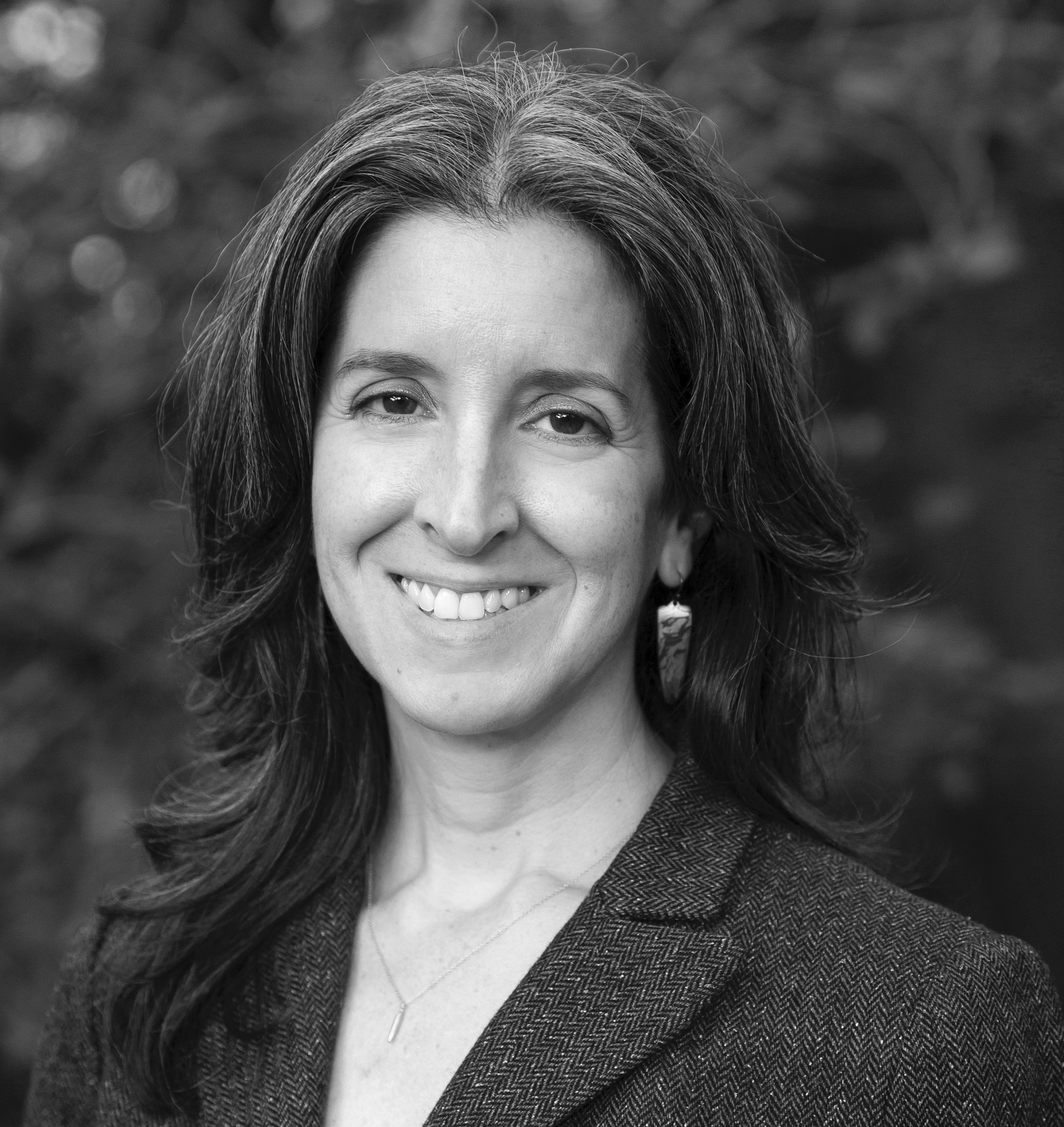 Black and white portrait of a smiling woman with long, wavy hair wearing a patterned blazer. She is standing outdoors with trees softly blurred in the background, exuding a timeless elegance that captures the spirit of Berkeley Journalism.