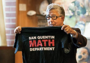 A man with gray hair and glasses, reminiscent of Bill Drummond, holds up a black T-shirt that reads "SAN QUENTIN MATH DEPARTMENT" in white and red letters. He stands indoors with various objects visible in the background.