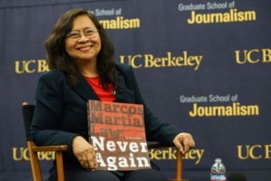 A woman with long dark hair, wearing glasses and a dark blazer over a red shirt, sits on a chair holding a book titled "Marcos Martial Law: Never Again." Behind her is a backdrop with text "Graduate School of Journalism UC Berkeley," highlighting the importance of Investigative Reporting.