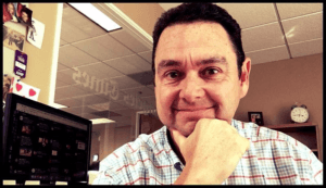 A man with short dark hair and a striped shirt is smiling at the camera while resting his chin on his hand. He is sitting in an office with a computer screen and a wall clock in the background. An alumni portrait hangs above photos pinned on a partition beside him.